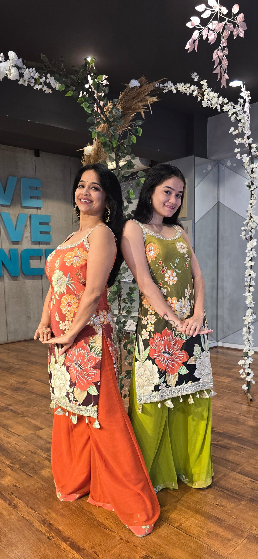 Two women in colorful floral dresses standing on a wooden floor with decorative elements.