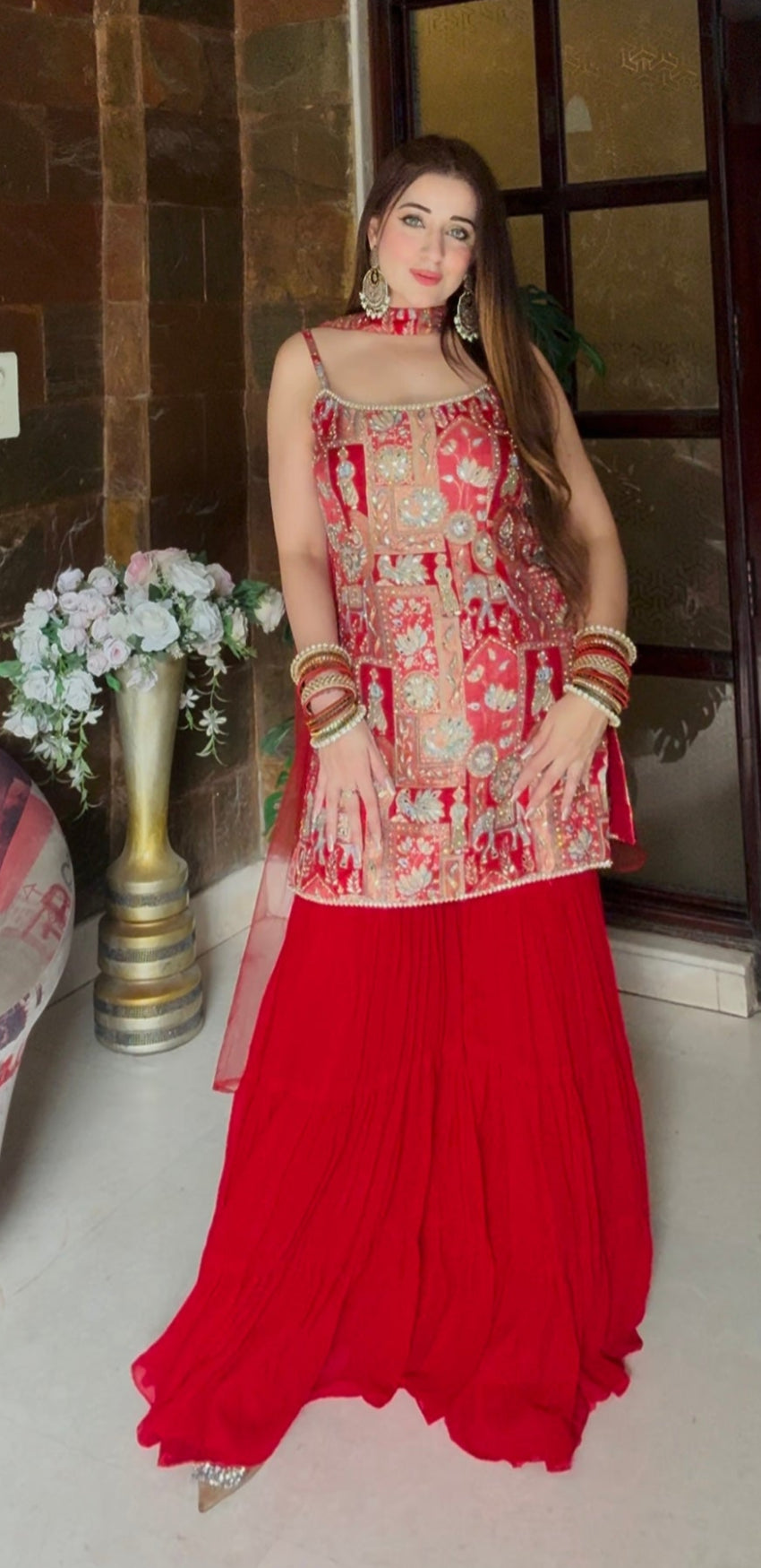 Woman in a red and gold traditional outfit standing in a room with decorative elements.