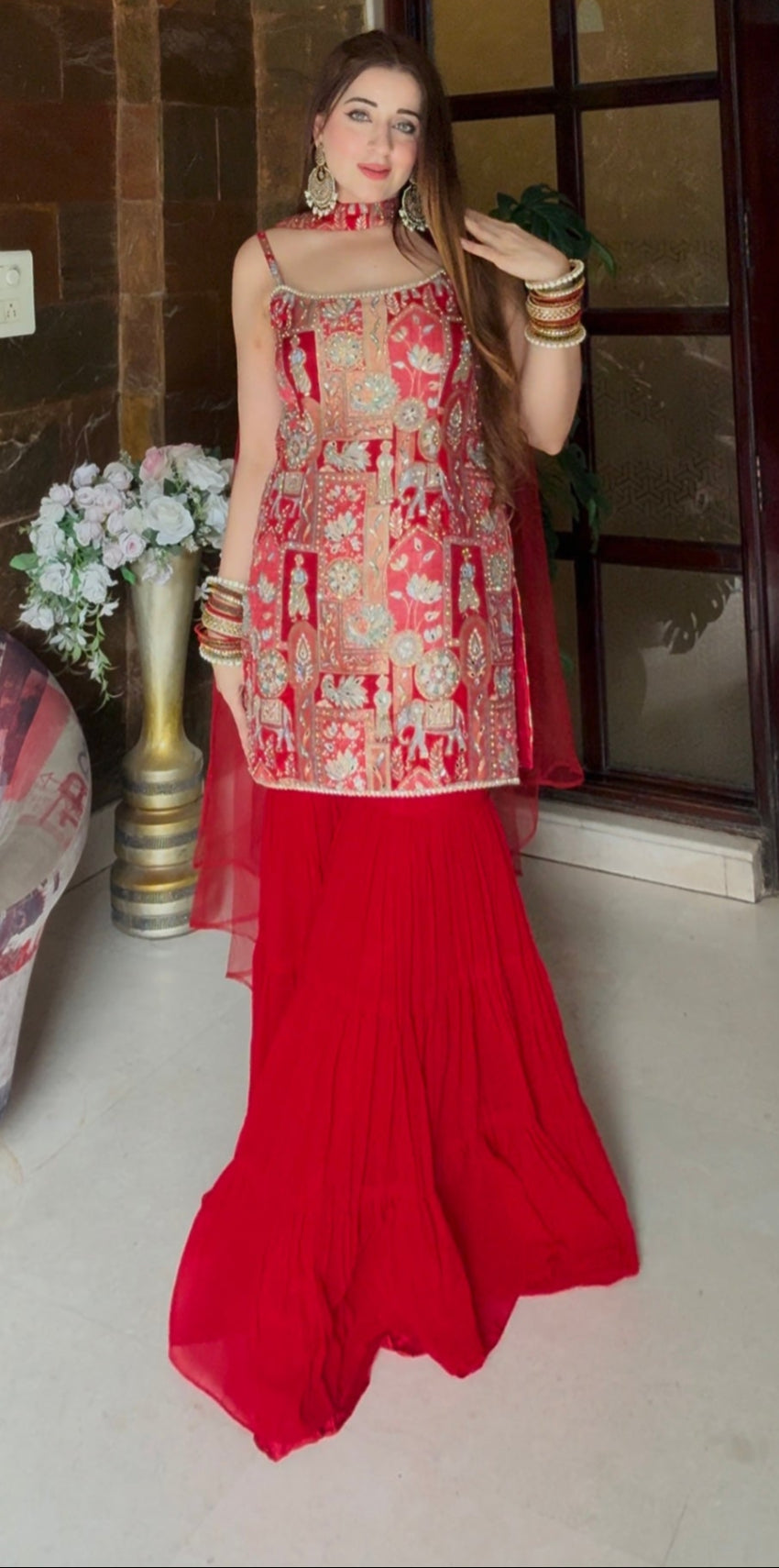 Woman in a red and gold traditional outfit standing in a room with marble walls and a decorative vase.