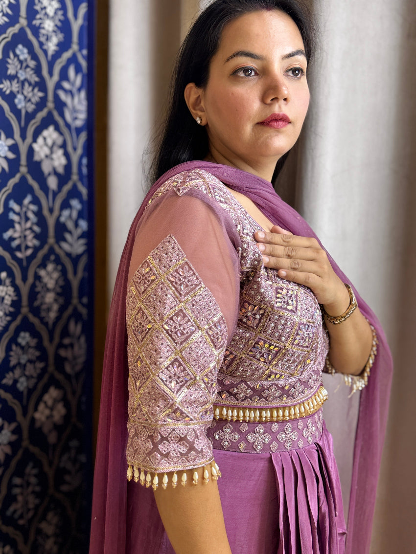 Woman wearing a traditional outfit with a purple blouse and pink dupatta against a decorative wall.