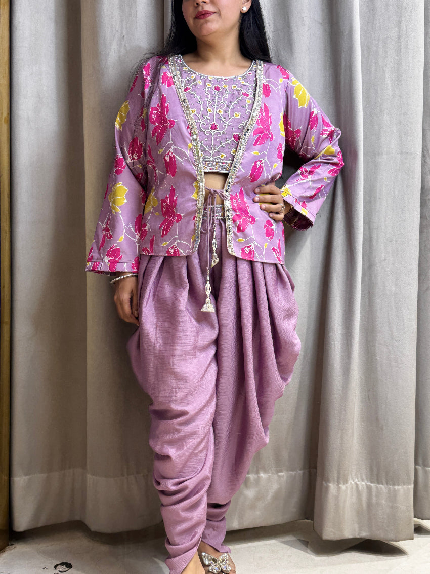 Person wearing a traditional outfit with a pink and floral top and purple dhoti against a neutral curtain background.