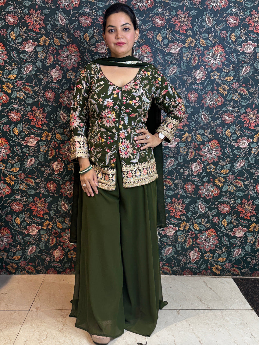 Woman in a green traditional outfit standing against a floral patterned wall.