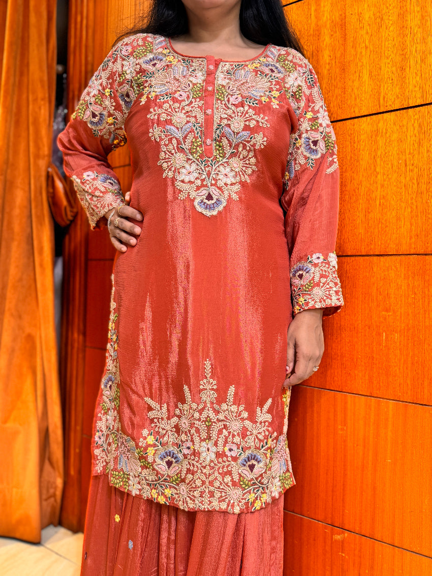 Person wearing a coral pink embroidered traditional outfit against an orange wall.