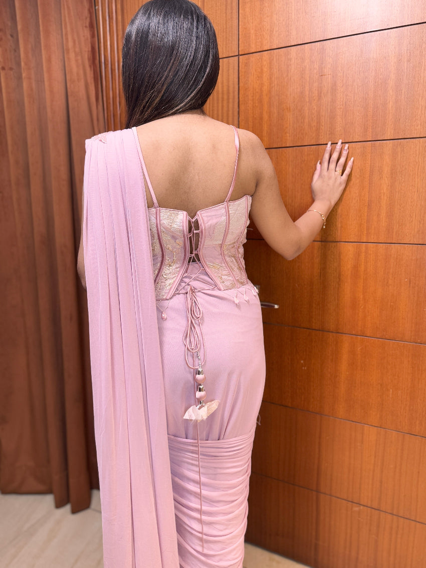 Woman in a pink saree standing against a wooden panel wall.