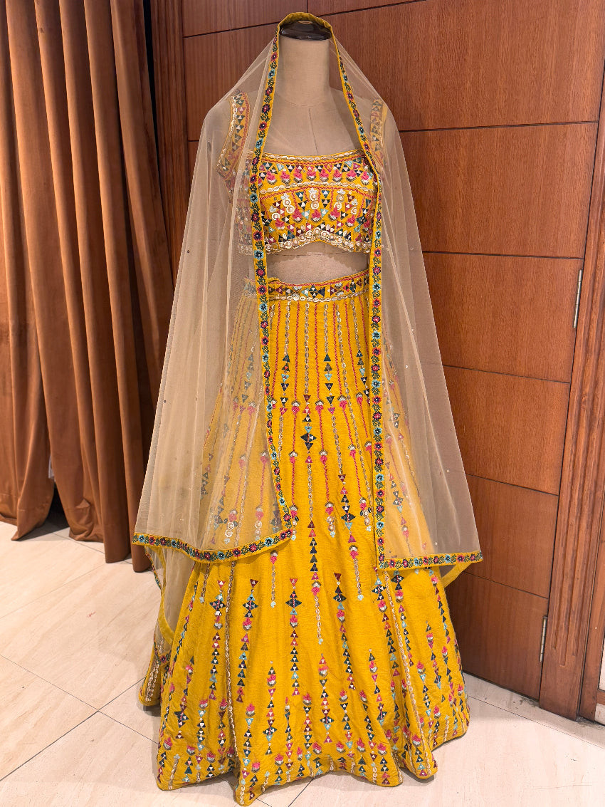 Yellow embroidered traditional outfit with a dupatta on a mannequin against a wooden panel background.