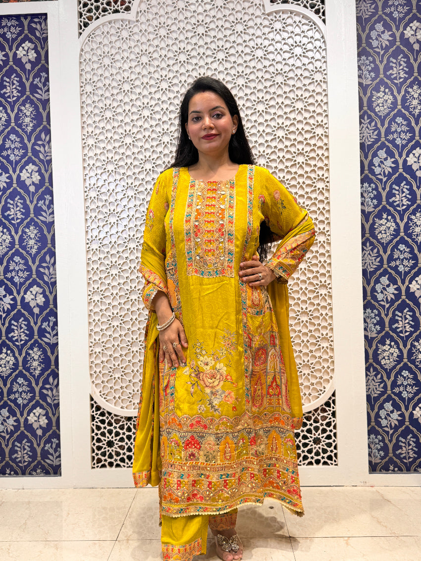 Woman in a yellow traditional outfit with intricate patterns standing against a decorative wall.