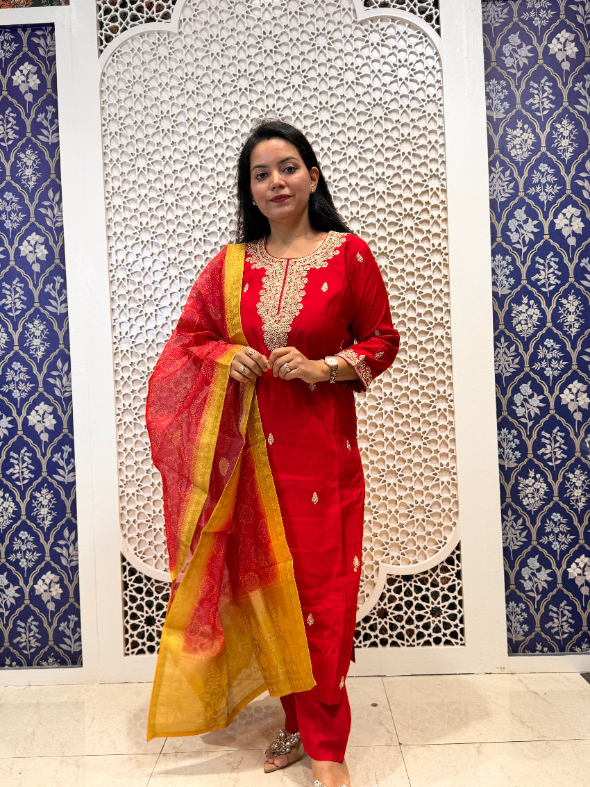 Woman in a red traditional outfit with yellow dupatta standing against a decorative wall.