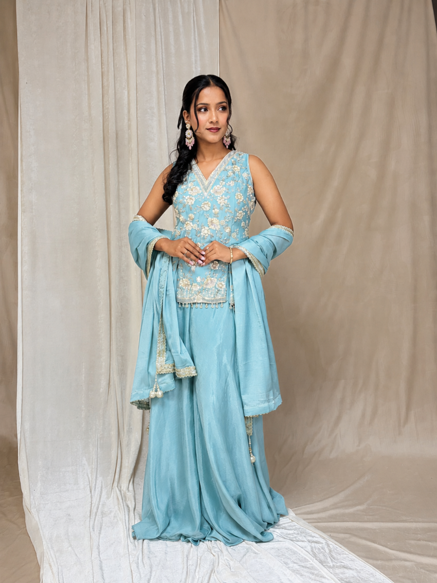 Woman wearing a light blue traditional outfit with floral patterns against a beige curtain background