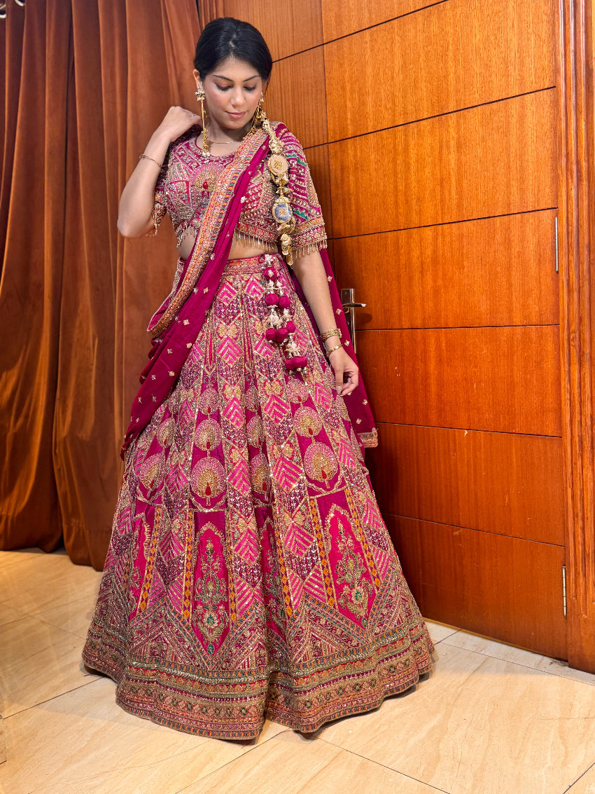 Woman in traditional pink and red embroidered lehenga standing against a wooden panel background