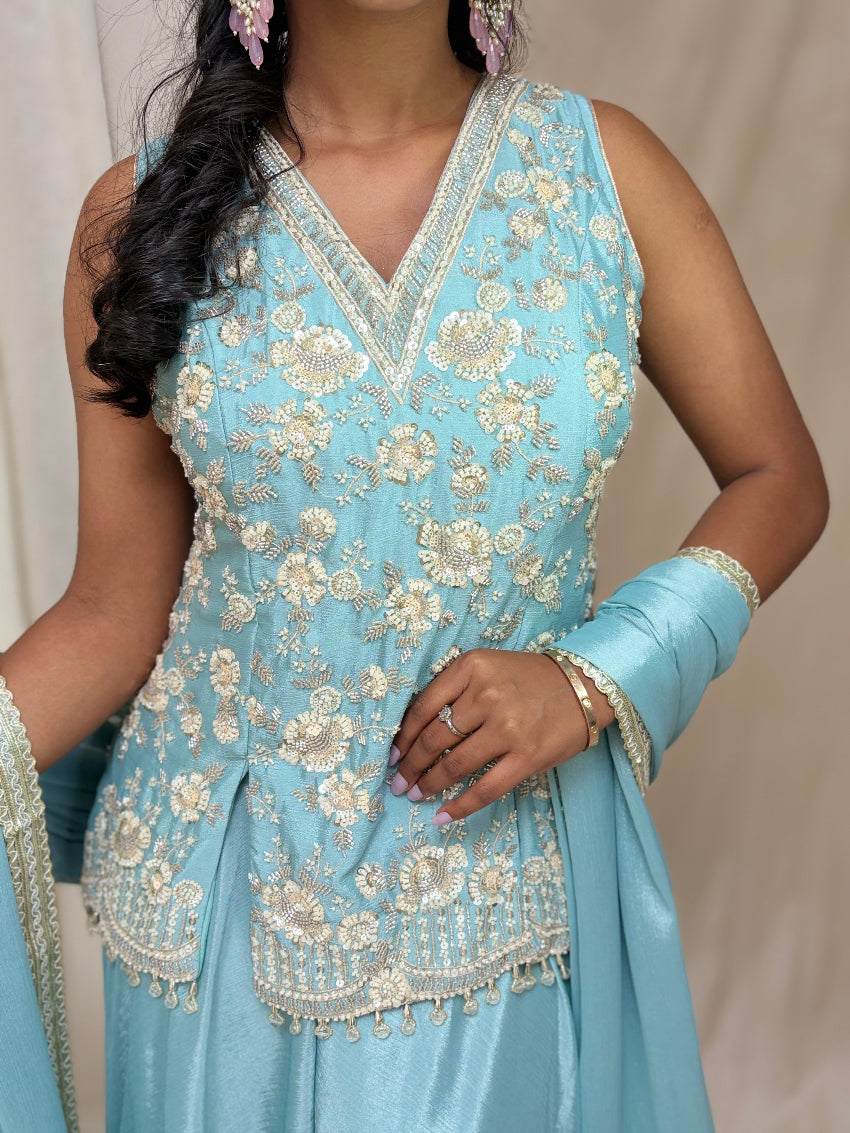 Woman wearing a light blue embroidered traditional outfit with matching gloves against a neutral background