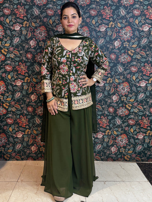Woman in a green traditional outfit standing against a floral patterned wall.