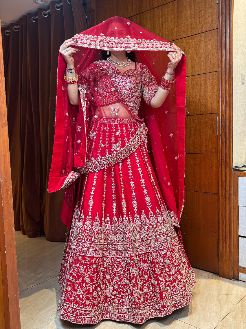 Red embroidered traditional outfit with a matching dupatta held by a person indoors.