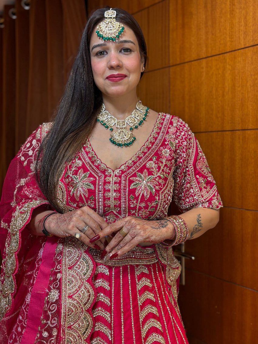 Woman in traditional red and gold embroidered outfit with jewelry against a wooden wall.