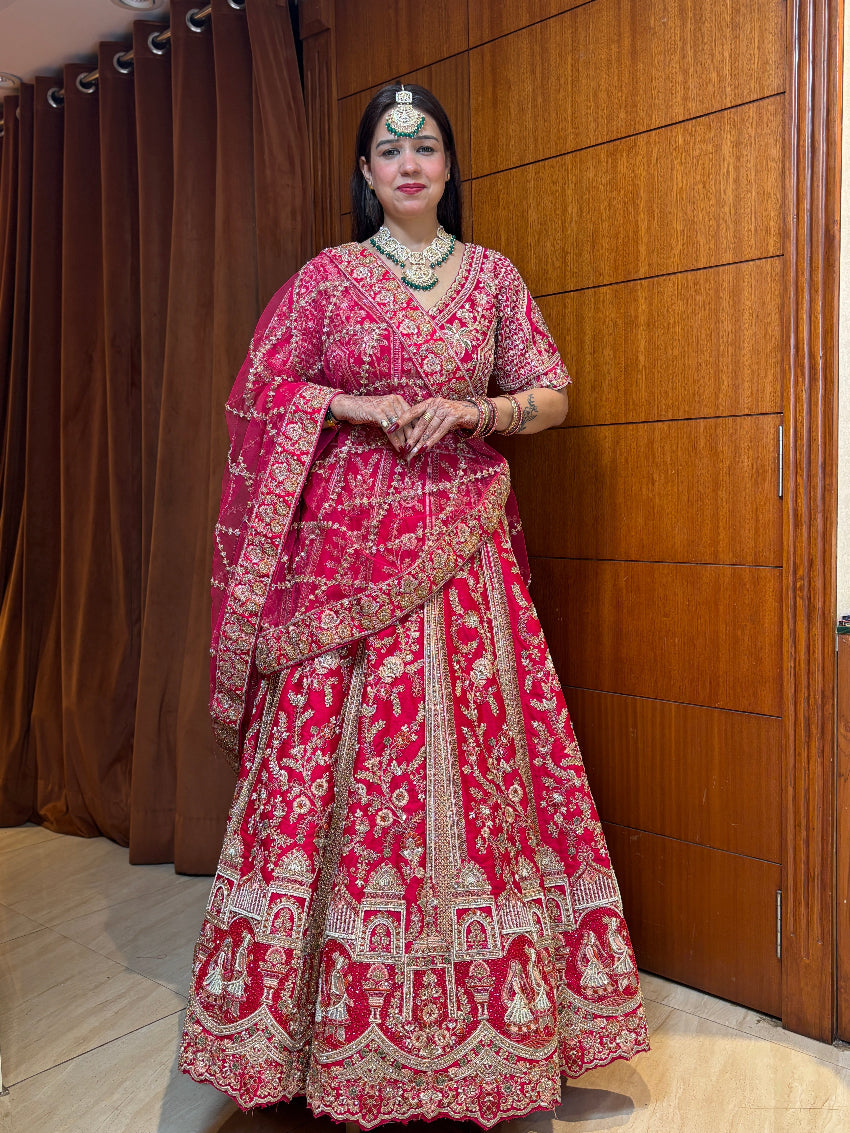 Woman in a traditional pink and red embroidered outfit standing against a wooden panel background.