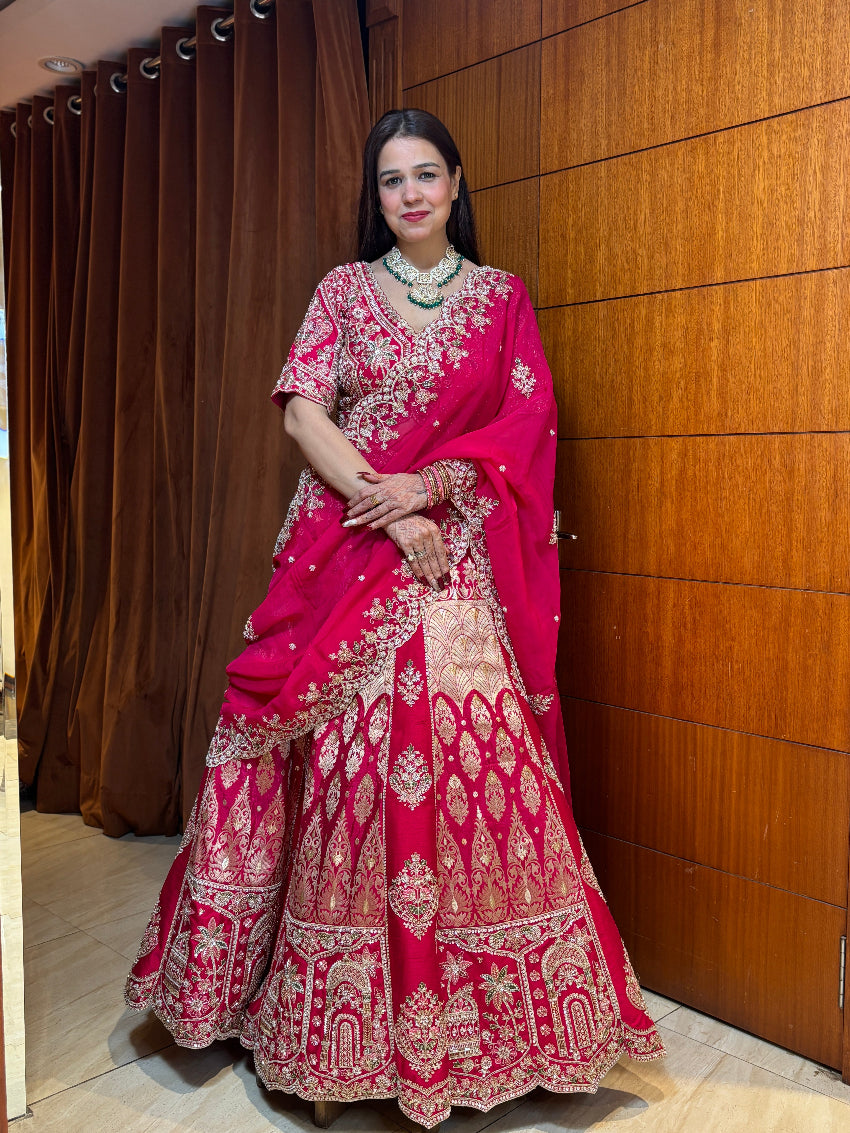 Woman in a traditional pink and red embroidered outfit standing against a wooden panel background.
