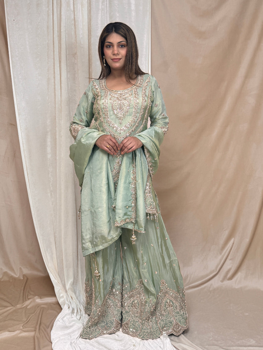 Woman wearing a light green embroidered traditional outfit against a beige curtain.