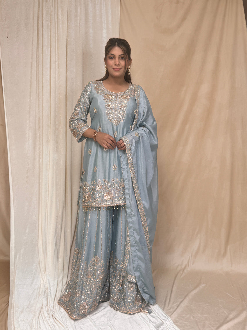 Woman wearing a light blue embroidered traditional outfit against a beige curtain.