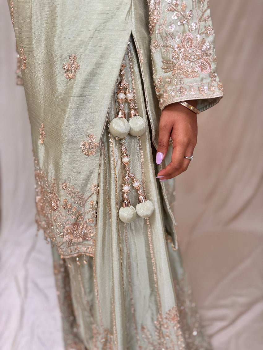 Light green embroidered traditional outfit with tassels and a hand holding a ring.