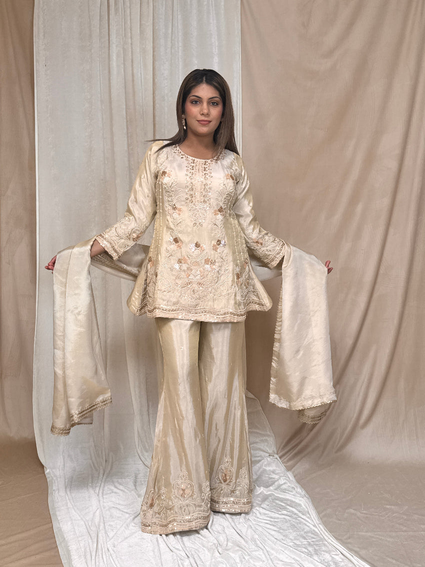 Woman wearing a traditional outfit with embroidered top and pants against a beige curtain background
