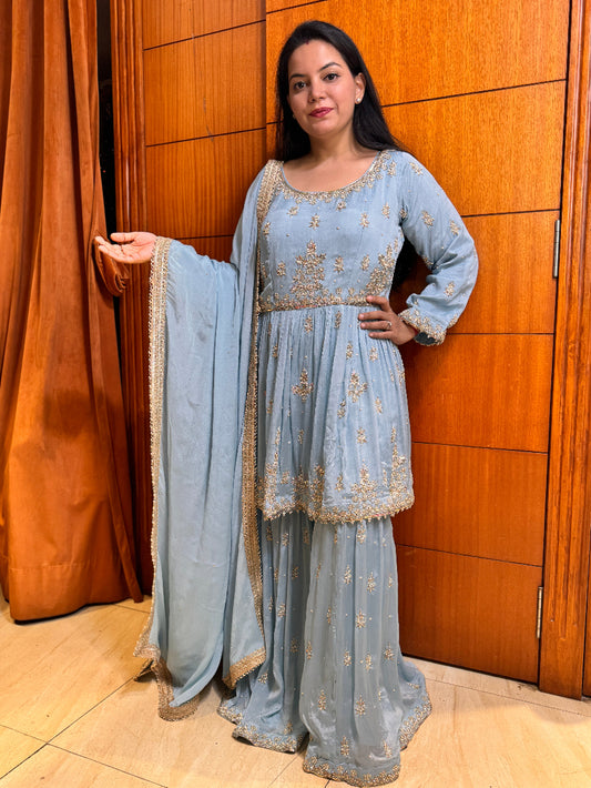 Woman in a light blue traditional outfit with gold embroidery standing against a wooden panel background.