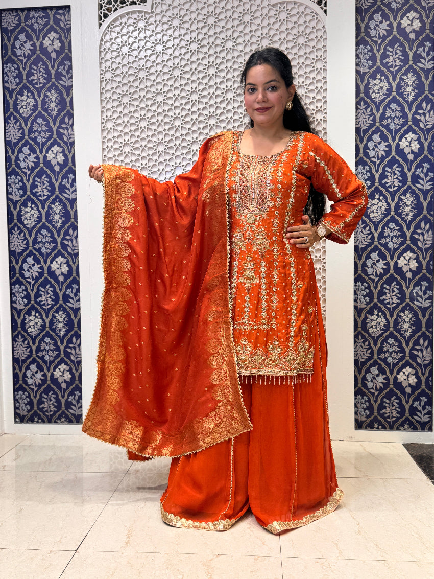 Woman in an orange traditional outfit with gold patterns against a decorative wall.