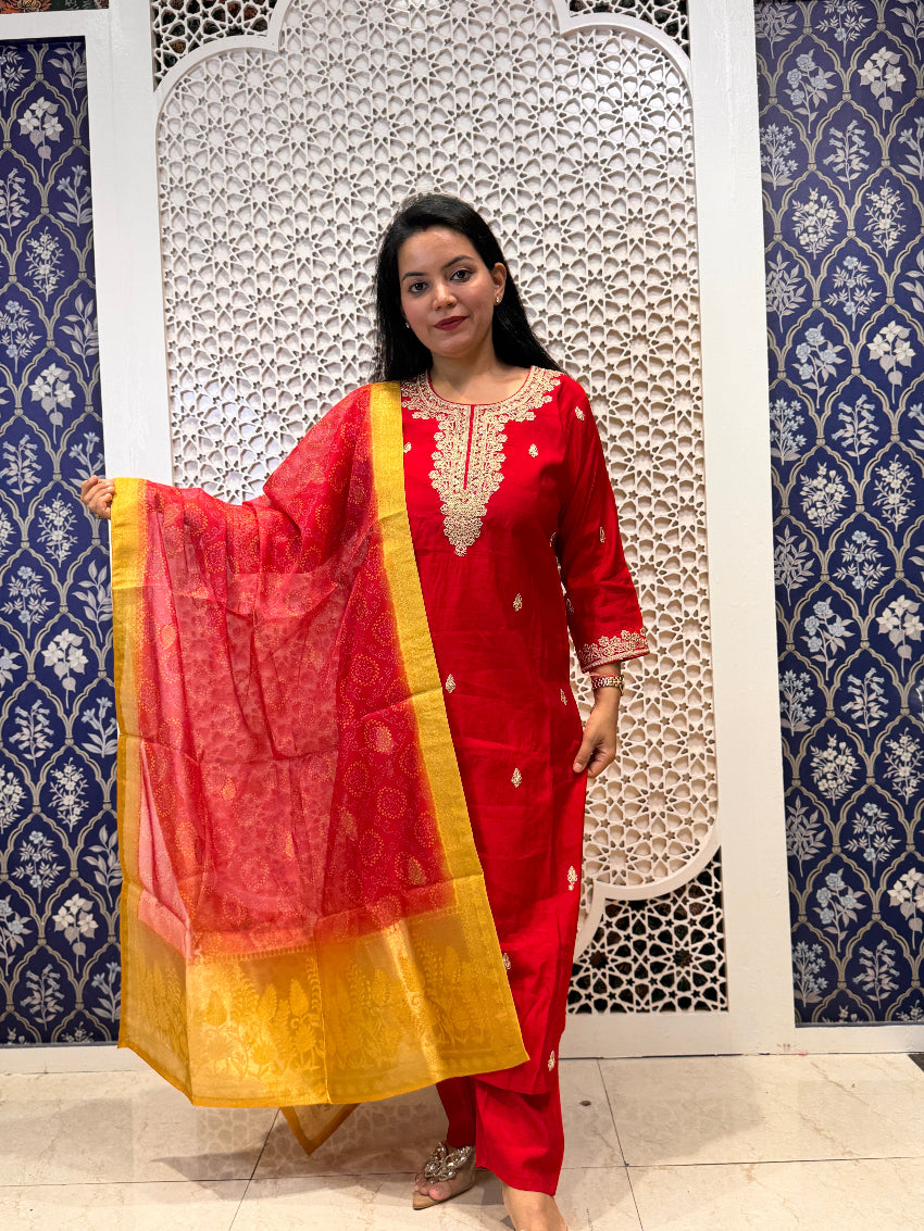 Woman in a red traditional outfit with a yellow dupatta against a decorative wall.