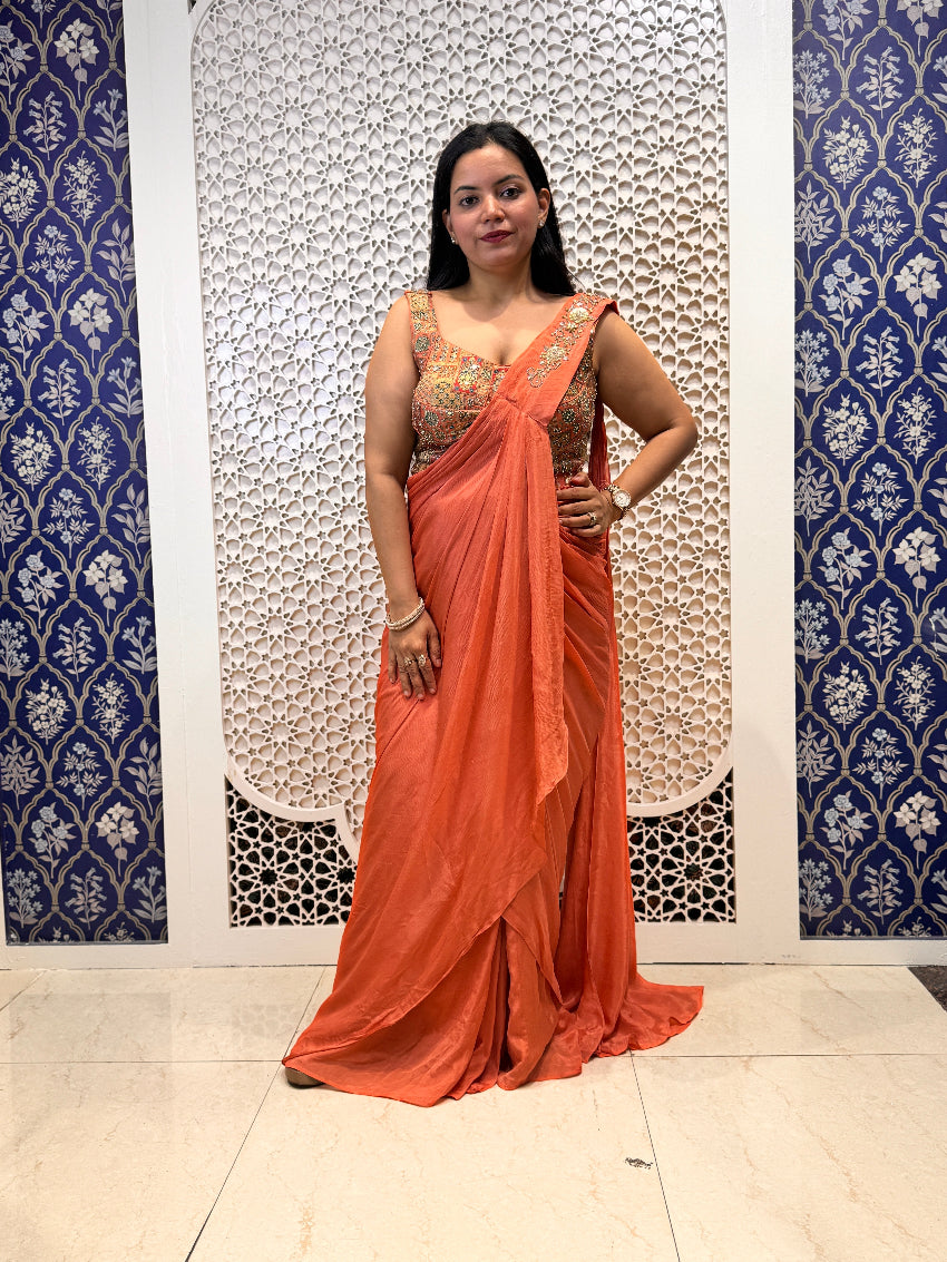 Woman in an orange saree standing against a decorative wall.