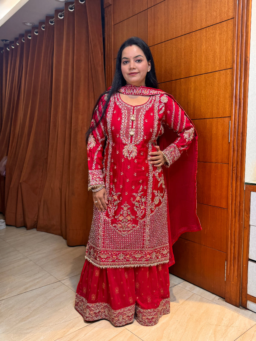 Woman in a red embroidered traditional outfit standing indoors against a wooden panel.