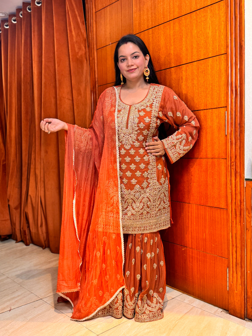 Woman in traditional orange and gold embroidered outfit standing against a wooden panel background