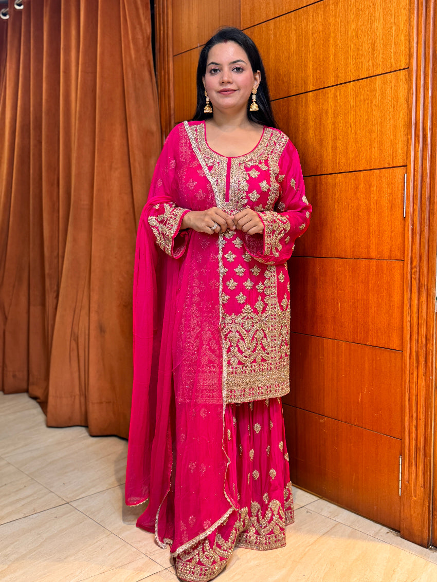Woman in a pink traditional outfit with gold embroidery standing against a wooden door.