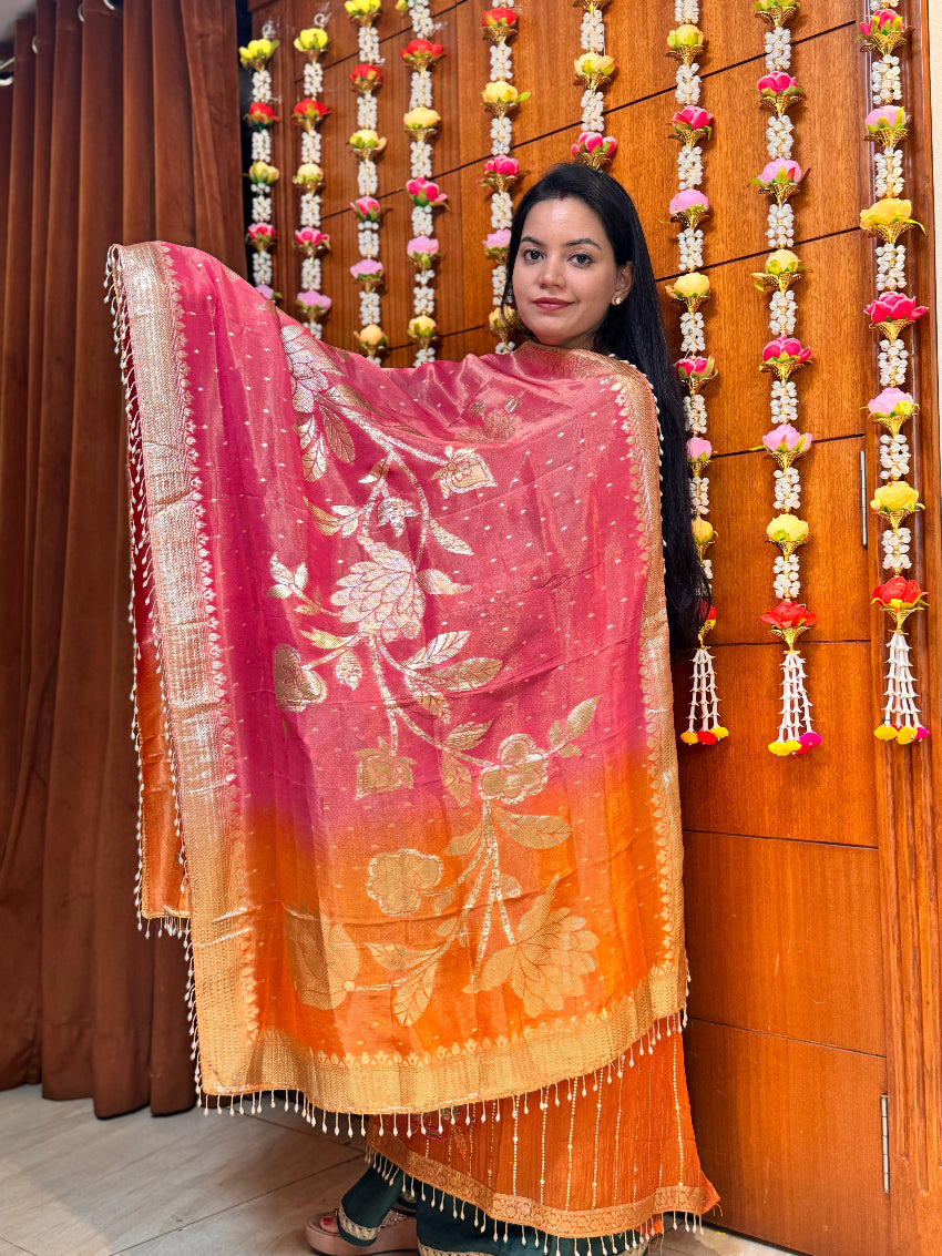 Woman wearing a pink and orange floral saree in front of a decorated wall.