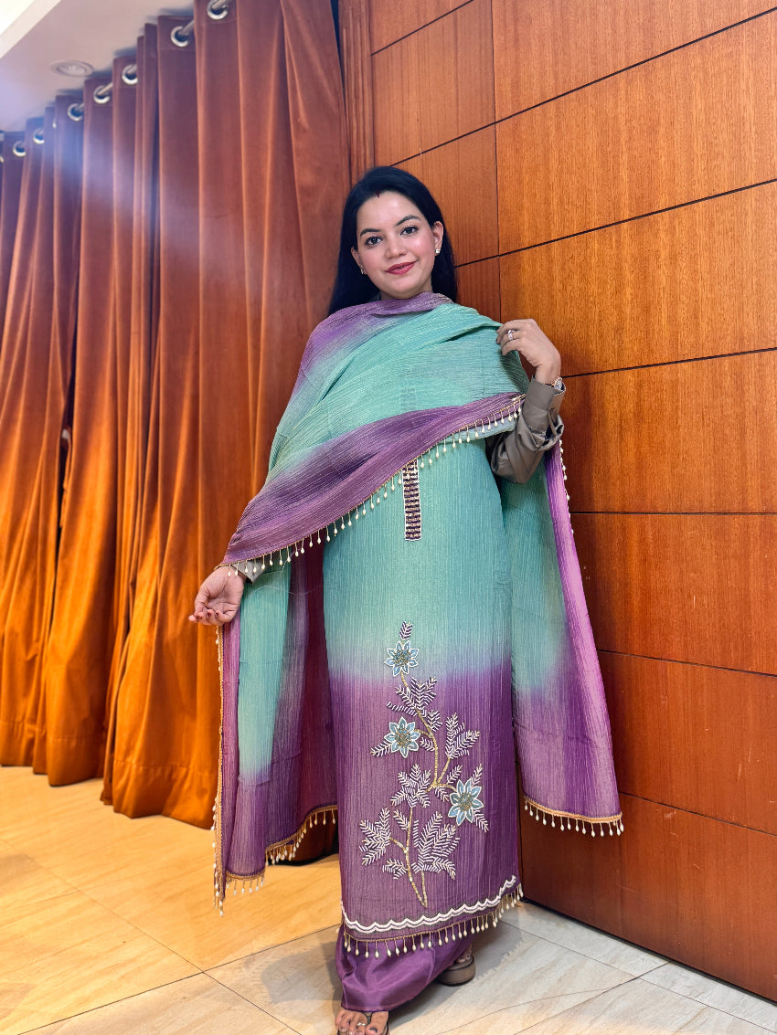 Woman wearing a traditional outfit with a purple and green dupatta against a wooden wall.