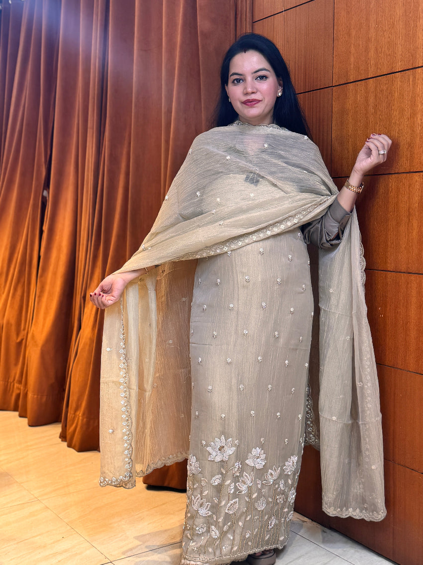 Woman in a beige embroidered outfit standing against a wooden wall.