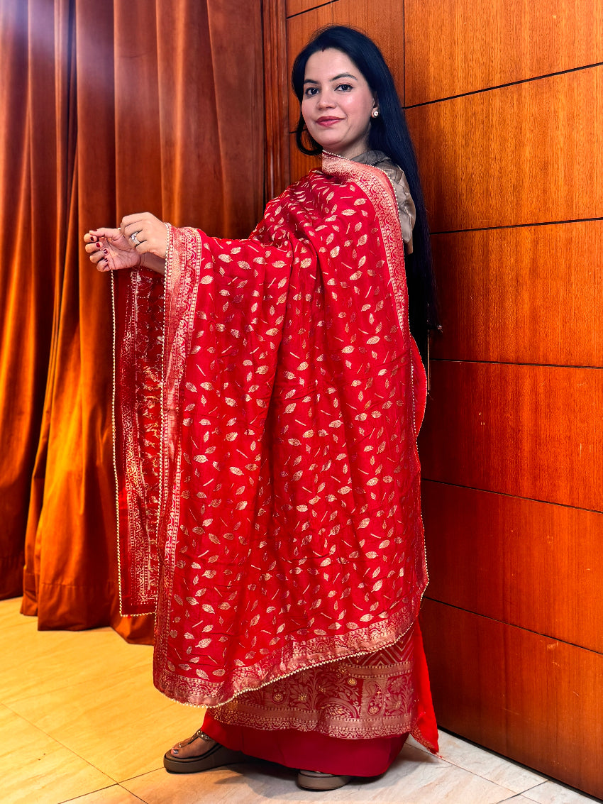 Woman wearing a red traditional outfit with gold patterns against a wooden wall.