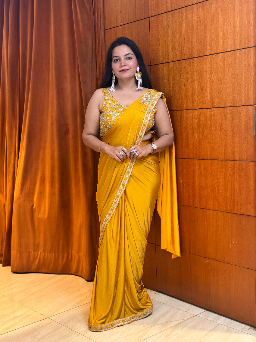 Woman in a yellow saree standing against a wooden paneled wall.