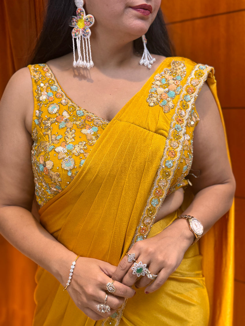 Woman wearing a yellow saree with intricate embroidery, holding it with both hands against an orange background.