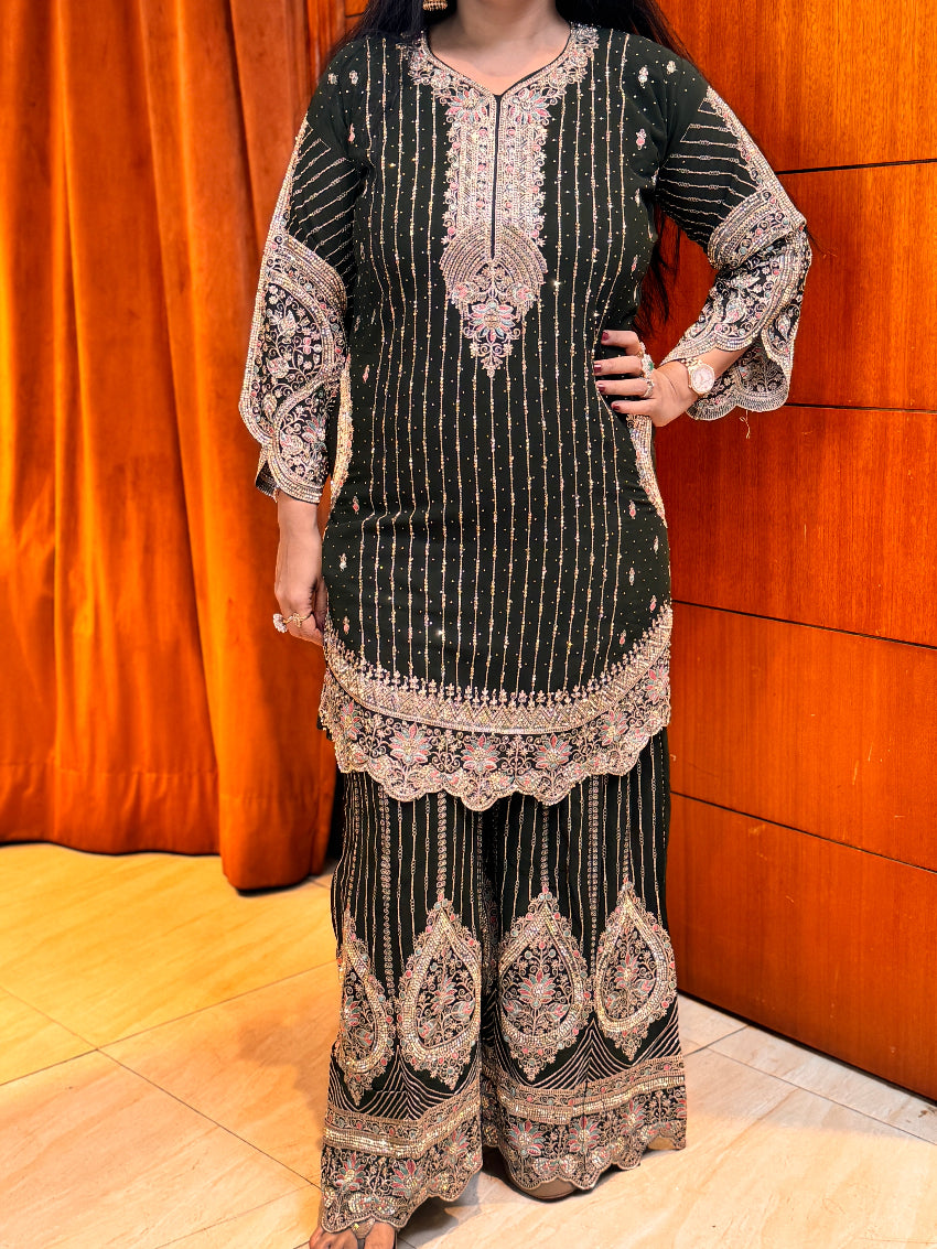 Person wearing a black and gold embroidered traditional outfit against a wooden panel background.