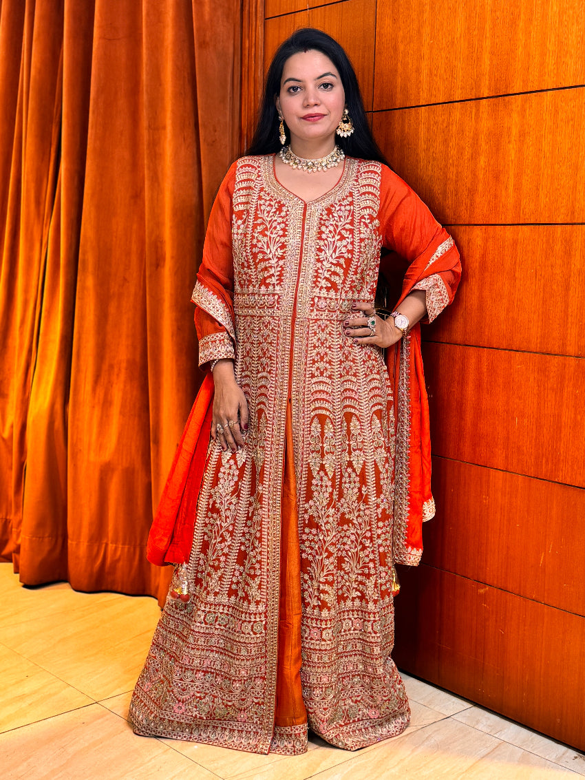 Woman in traditional red and gold embroidered outfit standing against a wooden wall.