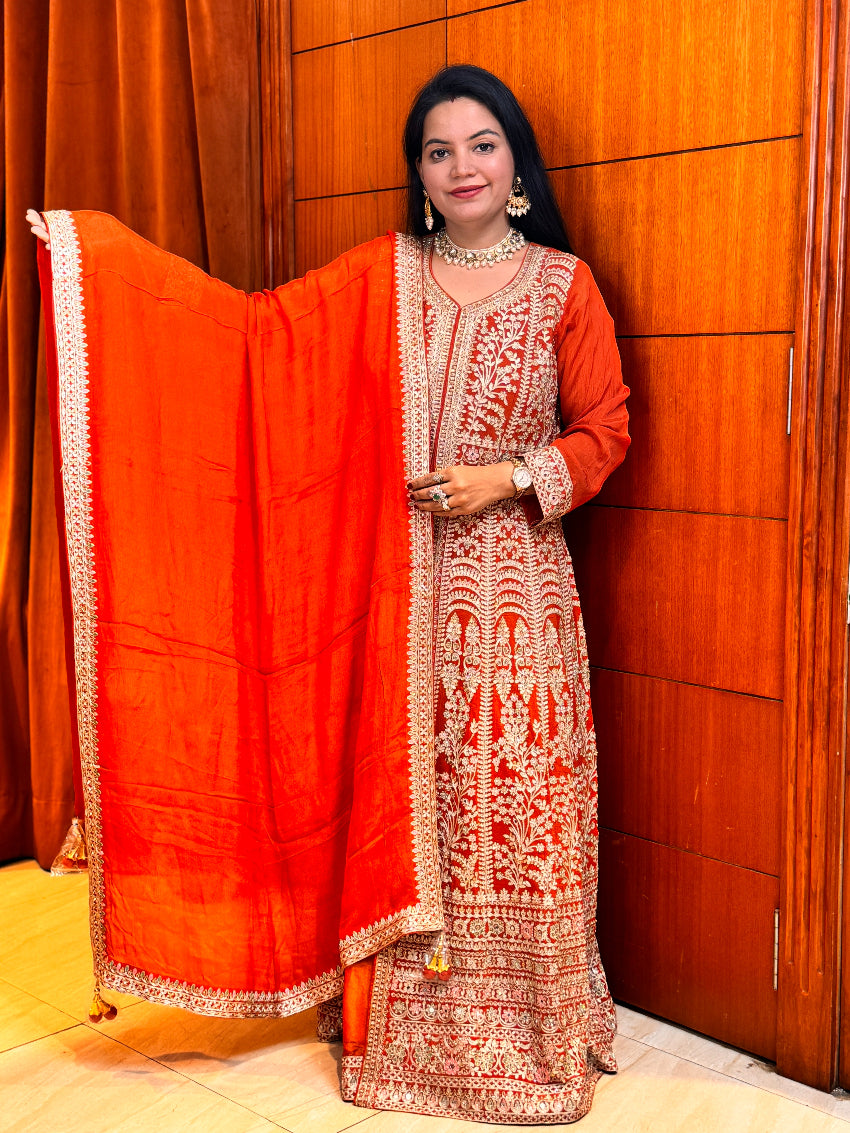 Woman in traditional orange and red embroidered outfit standing against a wooden wall.