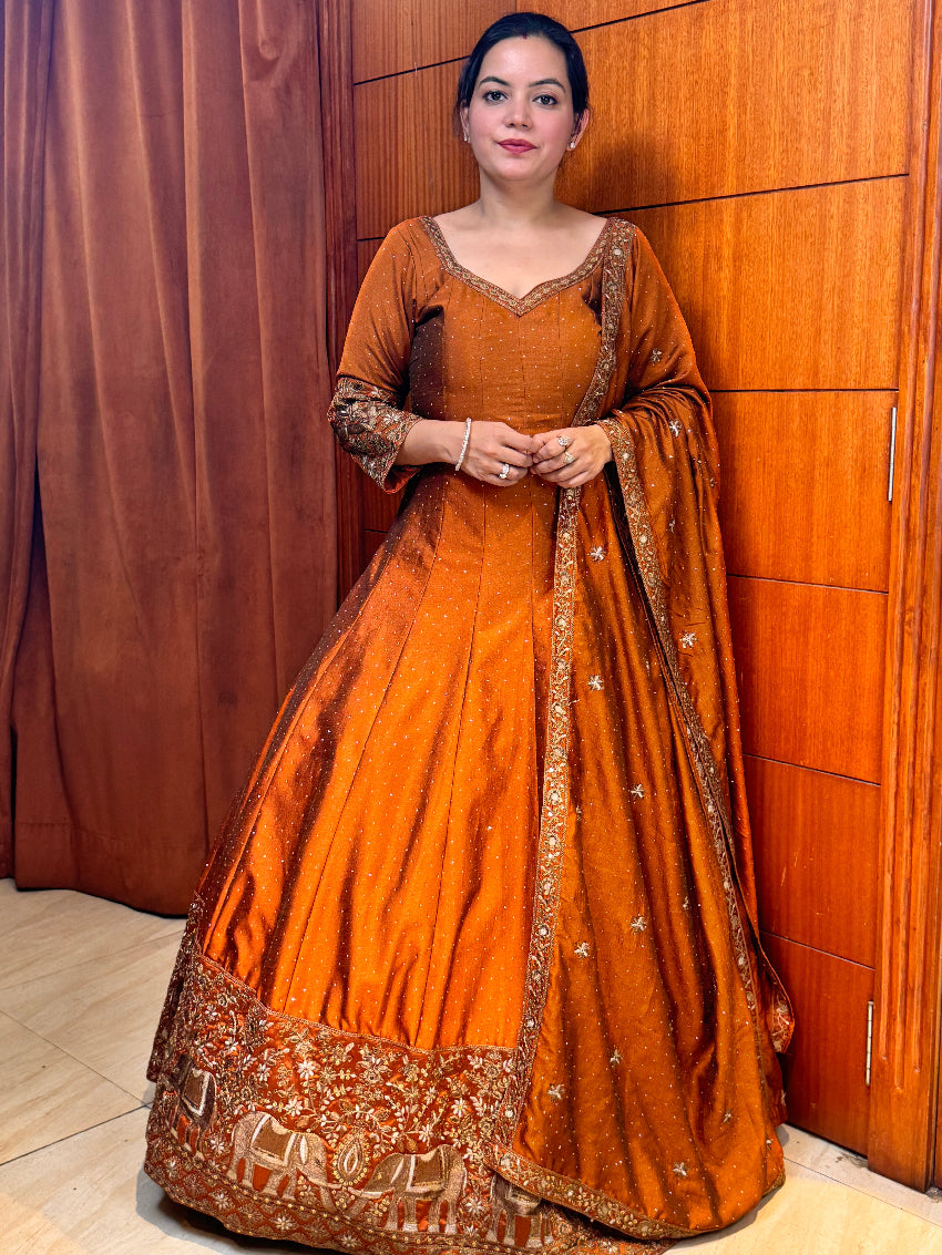 Woman wearing a traditional brown and gold outfit with intricate designs, standing against a wooden panel background.