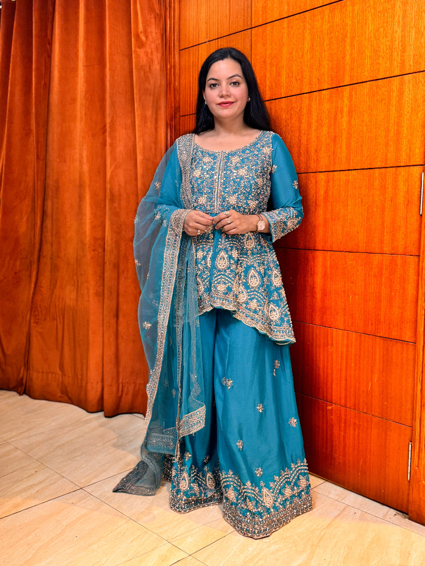 Woman in a blue embroidered traditional outfit standing against an orange curtain.