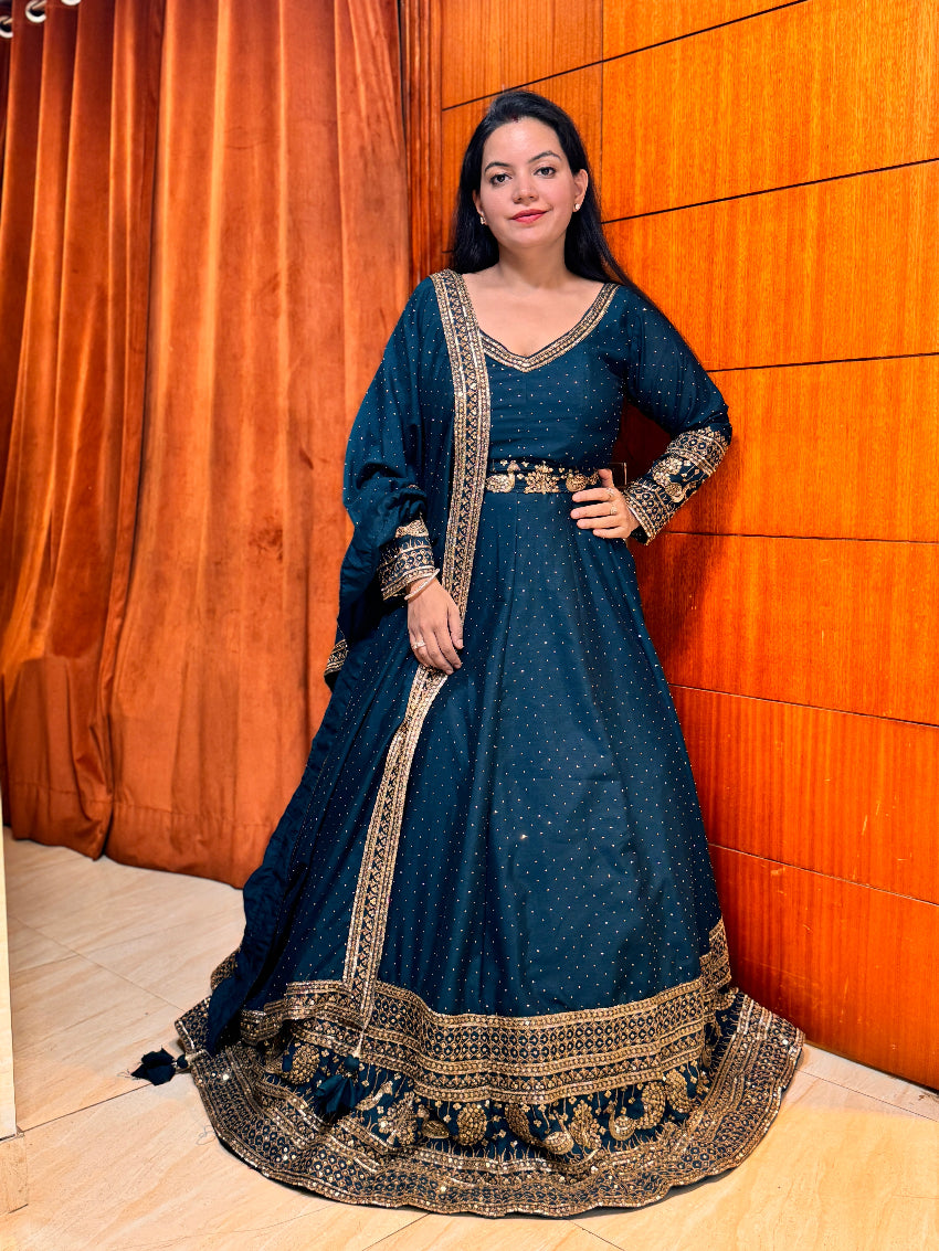 Woman in a blue traditional outfit with gold embroidery standing against an orange wall.