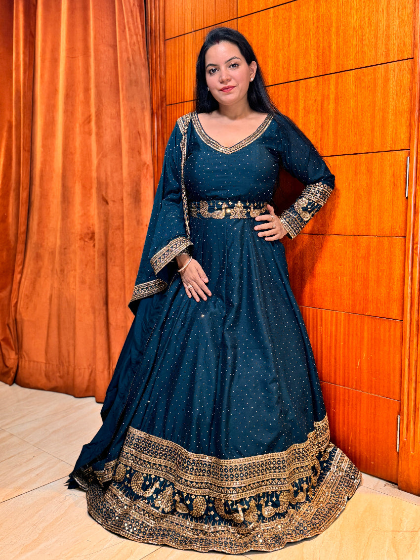 Woman in a dark blue traditional dress with gold embroidery standing against an orange wall.