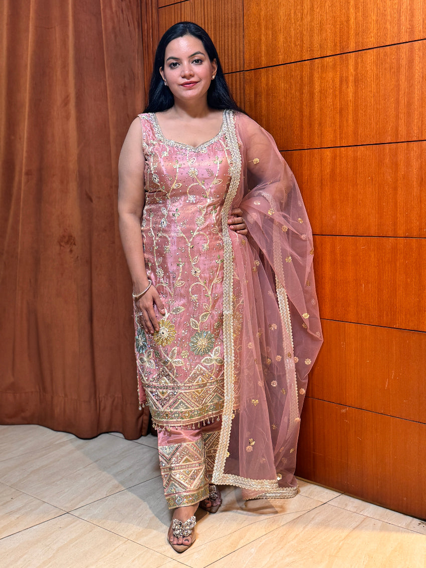Woman in a pink and gold traditional outfit standing against a wooden wall.