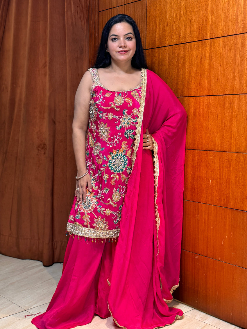 Woman in a pink traditional outfit with intricate designs against a wooden wall.