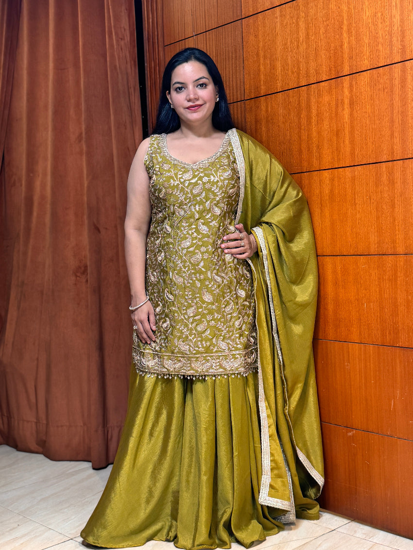 Woman in a green traditional outfit standing against a wooden paneled wall.