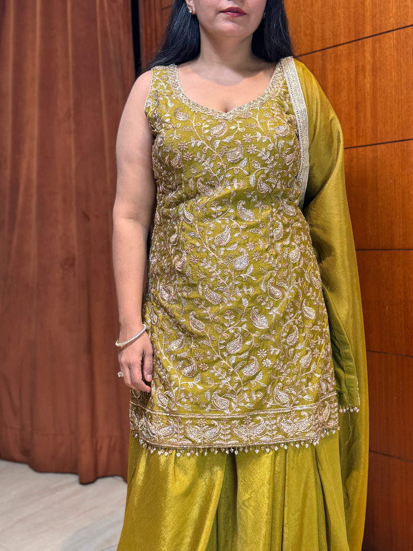 Woman wearing a green traditional outfit with intricate patterns against a brown curtain background