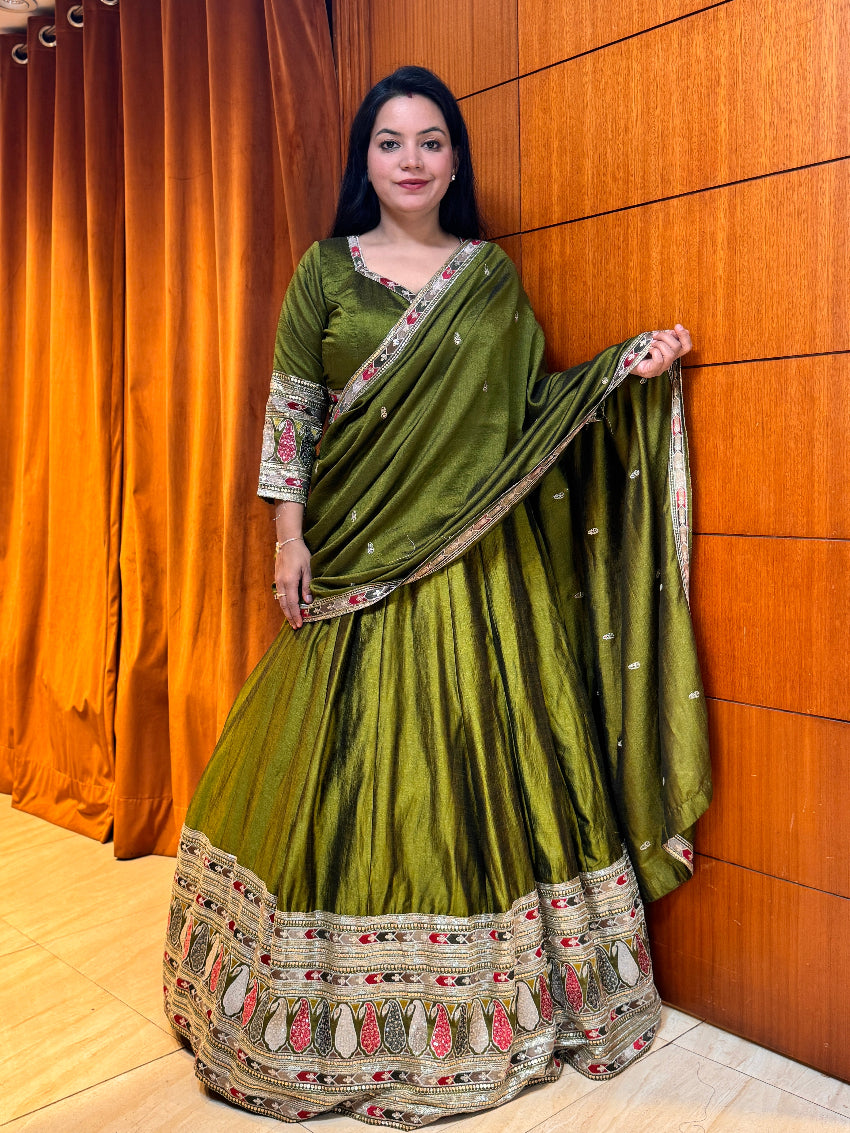 Woman wearing a green traditional outfit with intricate patterns against a wooden wall.