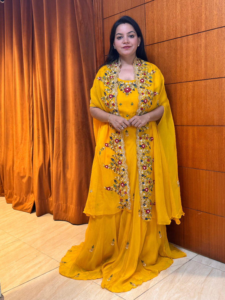Woman in a yellow traditional outfit with floral patterns standing against a wooden paneled wall.